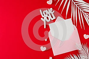 Flat lay photo of open envelope with card, white tropical leaves, hearts on red background
