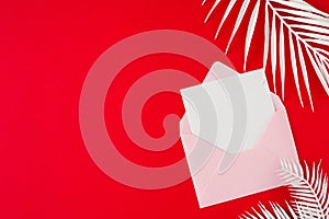 Flat lay photo of open envelope with card, white tropical leaves on red background