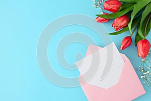 Flat lay photo of pink open envelope with white card, tulips and gypsophila flowers on pastel blue background