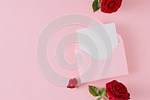 Flat lay photo of red flowers and open envelope with white card on pastel pink background