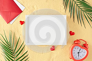 Flat lay photo of red hearts, green tropical leaves, alarm clock, envelope on sandy background and white frame