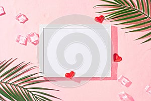 Flat lay photo of red hearts, tropical leaves, ice cubes on pink sandy background and white frame