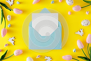 Flat lay photo of spring flowers, cute easter bunnies, white eggs on yellow background and envelope with letter