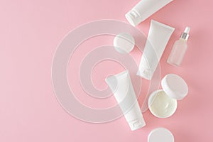 Flat lay photo of white cosmetic bottles, cream jars and dropper bottle on pastel pink background