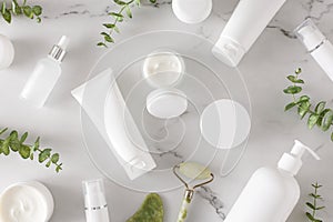 Flat lay photo of white cosmetic bottles, cream jars, dropper bottles, face massage roller and eucalyptus leaves