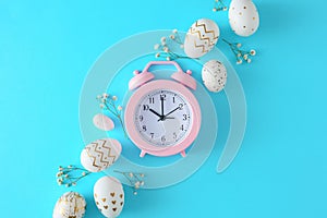 Flat lay photo of white gold eggs, gypsophila flowers on pastel blue background and pink alarm clock