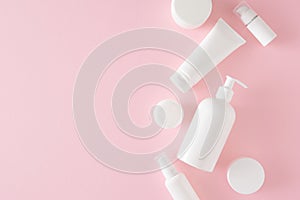 Flat lay photo of white pump bottle without label, cosmetic tubes and cream jars on pastel pink background