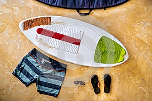 Flat lay photography of a white skimboard with red, white and green pads, with some orange surf wax on the nose in the middle of