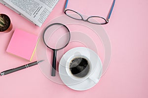 Flat lay on a pink background of coffee cup, glasses, magnifying glass, cactus, pen and newspaper