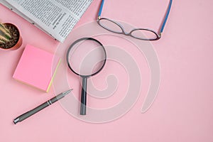 Flat lay on a pink background of glasses, magnifying glass, gadget, cactus, pen and morning newspaper