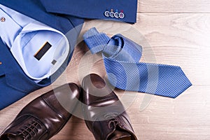 Flat lay set of classic mens clothes such as blue suit, shirts, brown shoes, belt and tie on wooden background.