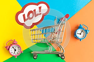 Flat lay of shopping cart and lol text in speech bubble with alarm clocks against colorful background minimal creative concept