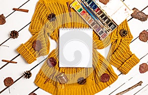 Flat lay of sketchbook, paints with brushes, yellow sweater, leaves, cinnamon sticks and pine cones on a white wooden background.