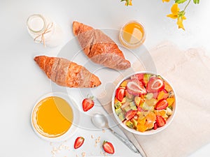 Flat lay summer breakfast with bowl of healthy fresh fruit salad, cup of tea and croissants on white background