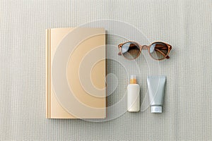 Flat lay of summer essentials including book, sunglasses, sunscreen and lotion on light textured surface, symbolizing