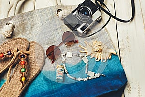 Flat lay of summer fashion with camera, slippers, sunglasses and other girl accessories on top of the bag on white wooden backgrou