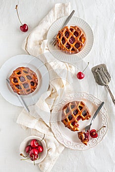 Flat lay of three cherry pies and fresh cherries on white plates  a white background and a serving utensil