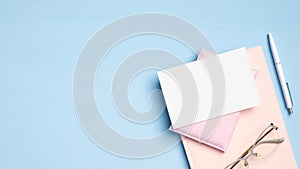 Flat lay, top view feminine workspace with pink notebook, glasses, document, pen and blank paper card on blue background. Modern