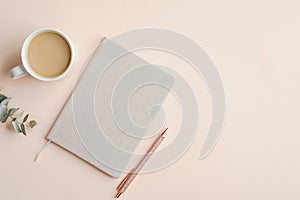 Flat lay, top view office table desk. Feminine workspace with paper notebook, pen, cup of coffee and eucalyptus leaf on beige