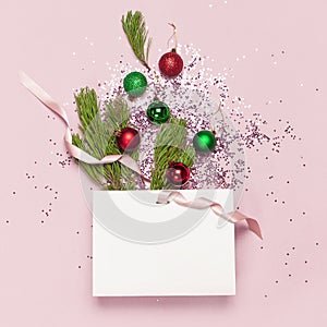 Flat lay top view White gift bag holographic glitter confetti red and green christmas balls green pine branches on pink background