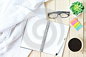 Flat lay or top view of white knitted blanket, open blank notebook paper, coffee cup and eyeglasses on wooden background