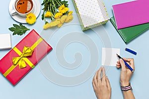 Flat lay top view womens hands write congratulations on empty sheet, red present box with gold ribbon, cup of tea, notebook, yello