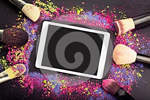 Flat lay of touchpad with blank screen with makeup brushes on colorful background of crumbled eyeshadows