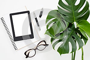 Flat lay tropical jungle Monstera leaves, paper notebook, e-book reader, glasses on white background. Top view feminine diary,