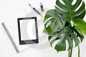 Flat lay tropical jungle Monstera leaves, paper notebook, e-book reader on white background. Top view feminine diary, stationery