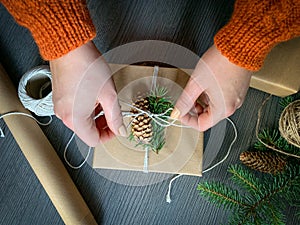 Flat lay of hands crafted gift box on wooden table with roll of craft paper, twine rolls, fir branches and pine cones photo