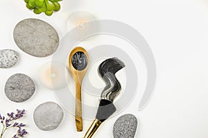 Flat lay view of mud charcoal mask on wooden spoon and smear on white background, surrounded with beauty brush and flat sea stones