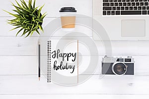 Flat lay view photo of working desk with happy holiday wish notebook, coffee cup, camera and laptop on white wooden background.