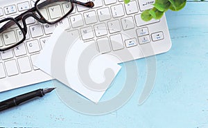 Flat lay white business card over keyboard with glasses, pen, tree on blue office desktop with copy space for text.