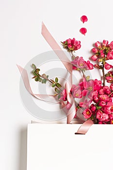 Flat lay White gift bag package with ribbon and spring pink flowers on light background top view. Greeting card with delicate