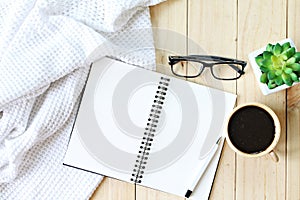 Flat lay of white knitted blanket, cup of coffee and blank notebook paper on wooden background