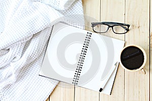 Flat lay of white knitted blanket, eyeglasses, cup of coffee and blank notebook paper on wooden background