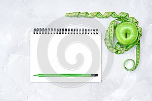 Flat lay of a whole green apple wrapped with a soft green measuring tape and a paper notebook with a pencil on a light gray concre