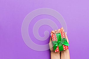 Flat lay of woman hands holding gift wrapped and decorated with bow on purple background with copy space. Christmas and holiday
