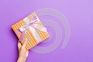 Flat lay of woman hands holding gift wrapped and decorated with bow on purple background with copy space. Christmas and holiday