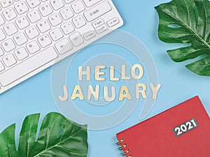 Flat lay of wooden letters HELLO JANUARY  with  red 2021 diary or planner, computer keyboard and monstera leaves  on blue