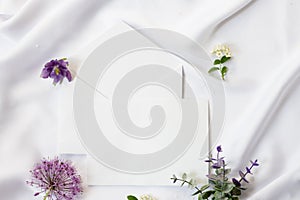 Flat lay workspace, mockup. Wedding invitation cards, craft envelopes, purple flowers, green leaves and lace on white