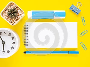 Flat lay, yellow office desktop with blank notebook and blue stationery, white clock, cactus. Top view with space for text.