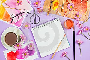 Flatlay with art supplies, artist palette, glasses, flowers, cup of tea and sketchbook with blank page