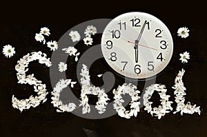 Flatlay big clock on a black background and the word school made of white petals. Concept back to school.