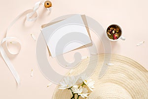 Flatlay composition with blank paper card mockup, envelope straw hat, daisy flowers, tea cup, ribbon on pastel pink background.