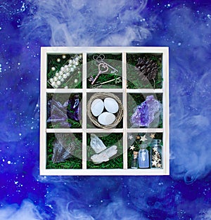 Flatlay of mysterious wooden witch box with moss and various things in it on a blue starry sky background with smoke