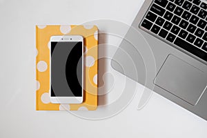 Flatlay of a white smarthpone, laptop and yellow dotted notebook, minimalist workspace