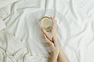 Flatlay of woman`s hands holding glass in lemon water in bed on white sheets