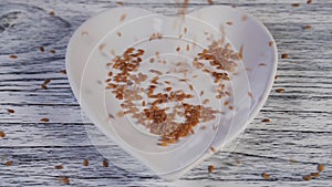 Flax seeds fall on a white heart-shaped plate on a light wooden table and bounce in different directions