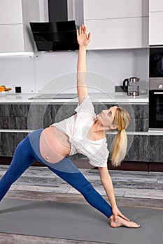 Flexible beautiful pregnant blond woman doing lunge exercise, standing in Extended Side Angle posture at home